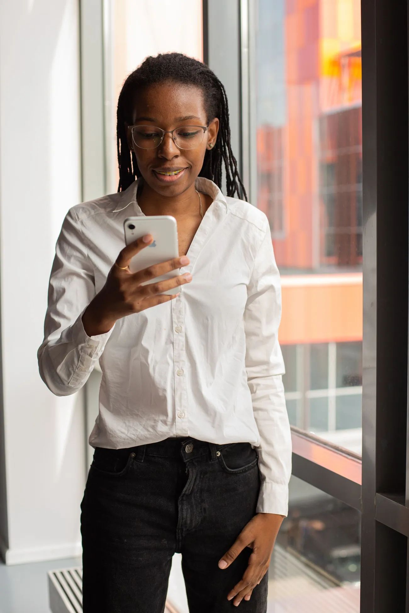 Uma mulher de camisa branca responde a uma mensagem no celular, de pé junto a uma janela